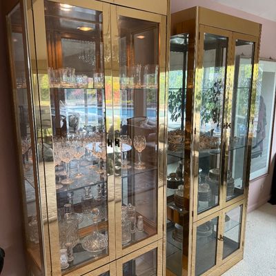 Pair of Hollywood Regency style brass-plated display cabinets with glass shelves, interior lighting, and mirrored backs, filled with...
