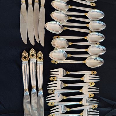 Vintage silver flatware set with gold-tone accents, including knives, spoons, and forks, displayed on a black background.