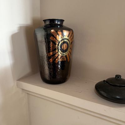 Hammered metal vase with a dark metallic finish, featuring a stylized copper and gold sunburst motif, displayed alongside a low black...