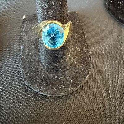 Gold ring featuring a large, oval-cut blue topaz gemstone in a bezel setting, displayed on a black velvet stand.
