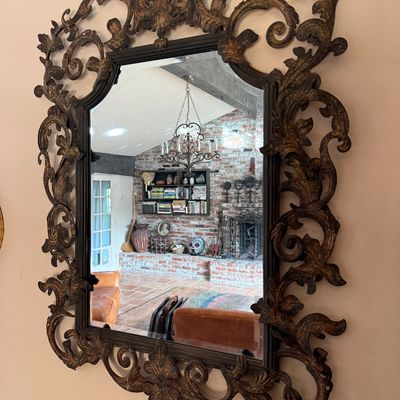 Ornate wrought iron wall mirror featuring a heavily detailed scroll and leaf frame with a distressed gold and dark bronze finish.