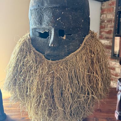 Hand-carved African wooden helmet mask featuring a dark, weathered finish and a full raffia beard, displayed on a metal stand.