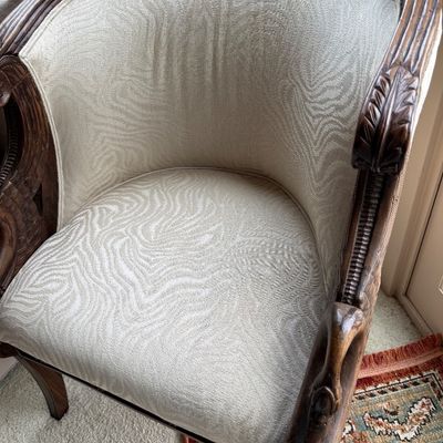 Vintage barrel-back armchair featuring carved dark wood trim and arms, upholstered in a textured cream-colored fabric with a subtle wavy...