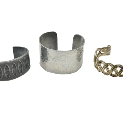 Trio of vintage metal cuff bracelets, featuring a dark grey patterned band, a wide hammered silver-tone cuff, and a braided wire design.