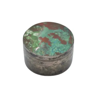 Small, round vintage pillbox featuring a tarnished silver-tone metal body and a lid inlaid with a colorful green, blue, and reddish-brown...
