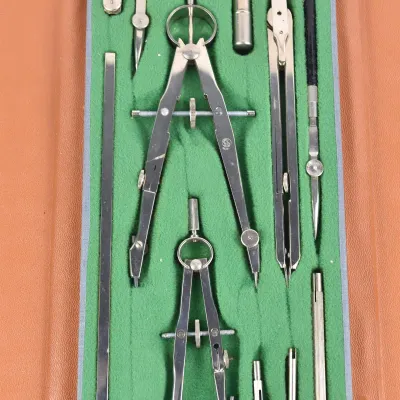 Vintage drafting tool set in a green felt-lined folding case, featuring various compasses, dividers, and ruling pens.