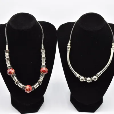 Two modern artisan necklaces on black velvet displays, one featuring ribbed silver-tone beads and red marbled spheres, and the other with...