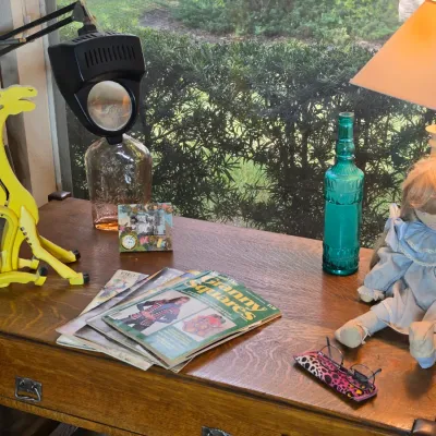 Vintage wooden desk featuring a collection of items including a yellow wooden giraffe, a magnifying desk lamp, vintage magazines, a teal...