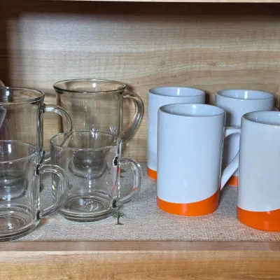 Assortment of glassware and mugs, including four clear glass coffee cups and four white ceramic mugs with orange bases, displayed on a...