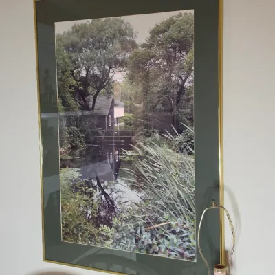 Framed landscape photograph depicting a rustic cabin by a reflective pond, presented in a dark green mat and a thin gold-toned metal frame.