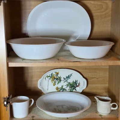 Assorted white ceramic and milk glass tableware, including serving bowls, platters, and small pitchers, displayed on wooden cabinet shelves.