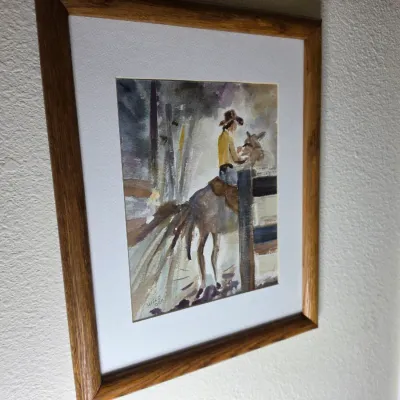 Framed watercolor painting depicting a cowboy on horseback, signed by the artist, in a wooden frame with white matting.