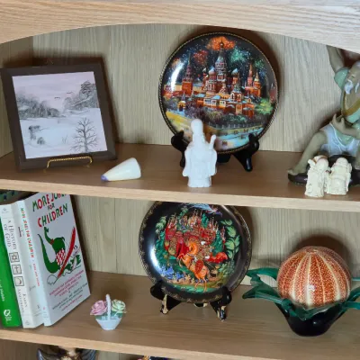Oak bookshelf displaying various collectibles, including Russian lacquer-style decorative plates, Asian figurines, a framed winter...