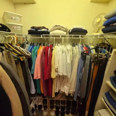 Walk-in closet filled with men's clothing, including hanging shirts, trousers, ties, belts, and folded sweaters on upper wire shelving.