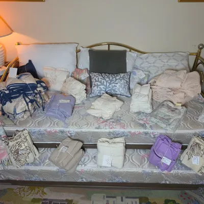 Brass daybed with trundle, featuring floral mattresses and a large assortment of folded linens, blankets, and decorative pillows.