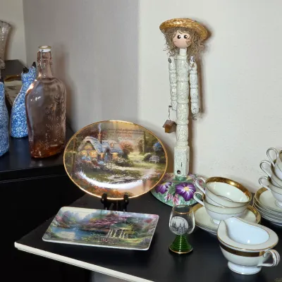 Assorted tabletop decor and tableware, including Thomas Kinkade collector plates, a whimsical wooden doll figure, blue and white ceramic...