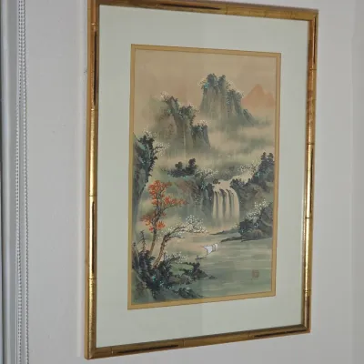 Framed Asian landscape painting depicting a waterfall, mountains, and a sailboat, housed in a gold-finished faux bamboo frame.