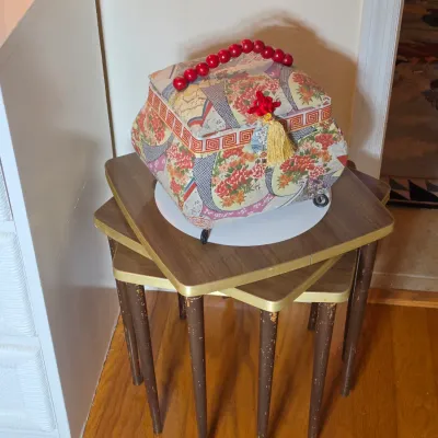 Vintage fabric-covered sewing basket with a red beaded handle and tassel detail, resting on a set of mid-century nesting tables with...