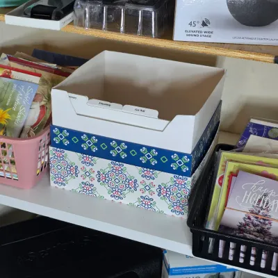 Assorted greeting cards, calendars, and organizational storage boxes arranged on a white shelf.