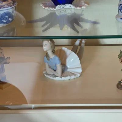 Collection of small figurines displayed in a glass cabinet, featuring a Lladro-style porcelain ballerina lying down, a small child...