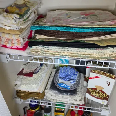 Assorted household linens, including placemats, hand towels, and lace doilies, organized on wire closet shelving alongside kitchen items.