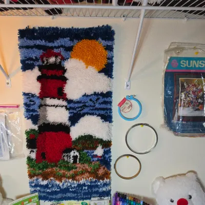 Vintage latch hook wall hanging featuring a lighthouse scene, displayed alongside embroidery hoops and craft kits.