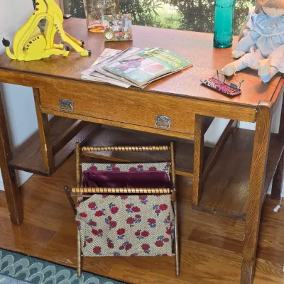 Vintage oak writing desk featuring a single drawer with metal pulls and lower side shelves, paired with a folding fabric magazine rack.