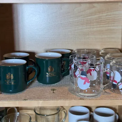 Collection of mugs on a cabinet shelf, including dark green mugs with gold crests and clear glass mugs featuring snowman illustrations.
