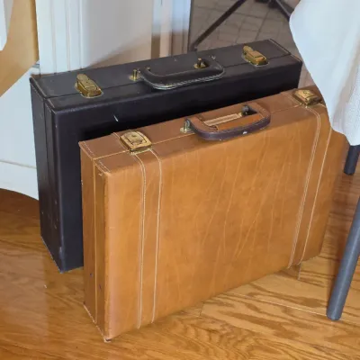 Two vintage hard-sided briefcases, one in black leather and one in tan textured vinyl, both featuring brass-toned latches and top handles.