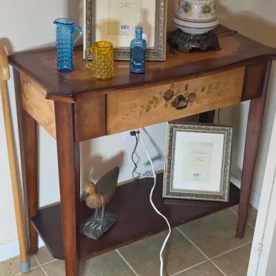 Two-tier wooden console table featuring a hand-painted floral drawer front and inlaid top, styled with vintage glassware, picture frames,...