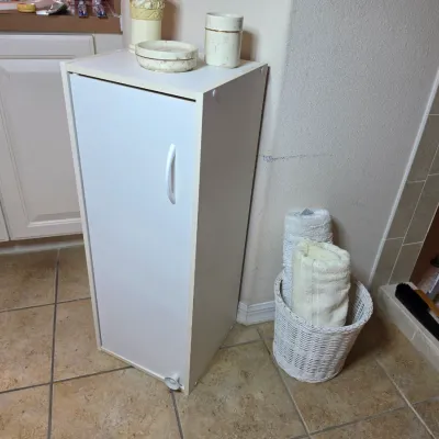 White laminate single-door bathroom storage cabinet, shown with a small white wicker basket holding rolled towels.