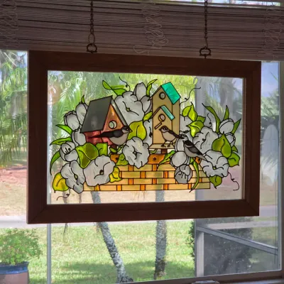 Stained glass window panel featuring a birdhouse and floral design, housed in a simple wooden frame and suspended by chains.