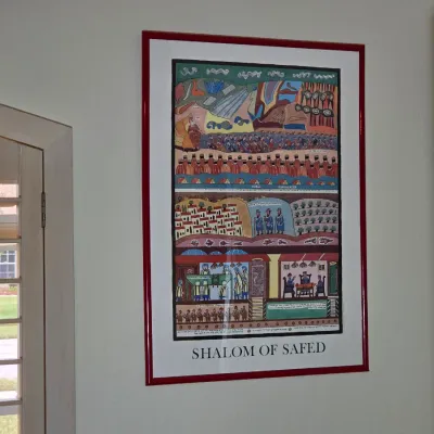 Framed "Shalom of Safed" art poster featuring colorful, detailed biblical and historical scenes, housed in a simple red frame.