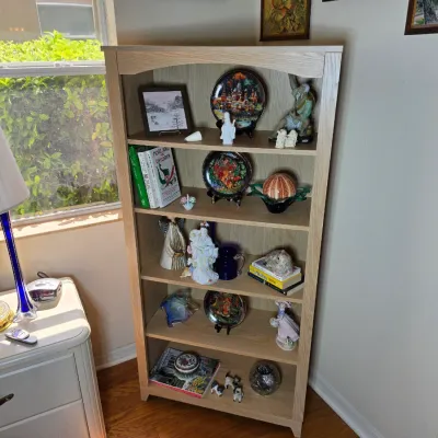 Light oak five-shelf bookcase displaying various decorative plates, figurines, books, and glassware.