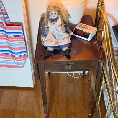 Vintage mahogany drop-leaf side table with a single drawer and inlaid details, displaying a porcelain doll and a RadioShack digital alarm...