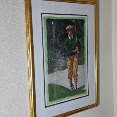 Framed watercolor painting depicting a golfer in vintage attire playing out of a sand trap, presented in a gold-finished frame with a...