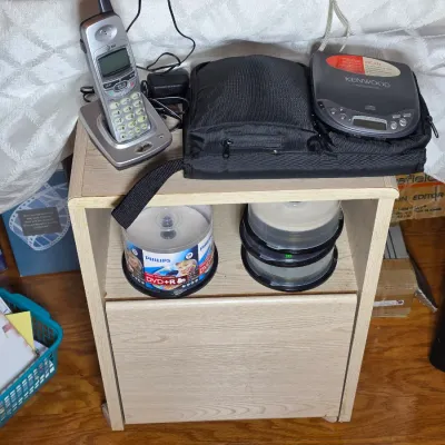 Light wood laminate rolling cart holding a Kenwood portable CD player, an AT&T cordless phone, and spindles of blank Philips DVD+R...