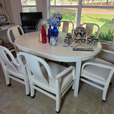 White oval dining table with six matching cushioned armchairs, styled with decorative glassware, blue hydrangeas, and a vintage ceramic...