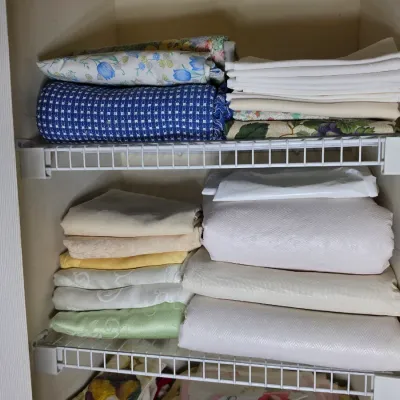 Assorted vintage bed linens and tablecloths, including floral, patterned, and solid white textiles, neatly folded on wire closet shelving.
