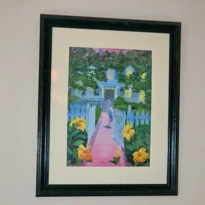 Framed and matted art print depicting a southern-style home with a white picket fence, yellow hibiscus flowers, and a heron on the path.