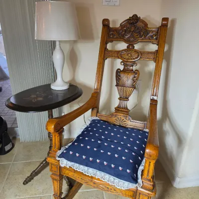 Ornate carved oak rocking chair with a blue patterned seat cushion, paired with a small oval side table and a white table lamp.