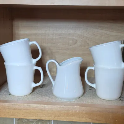 Vintage white milk glass coffee mugs and matching creamer pitcher, displayed inside a wooden cabinet.