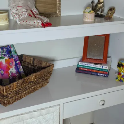 White wooden desk with hutch, featuring a woven storage basket, a stack of books with a picture frame, and various decorative...