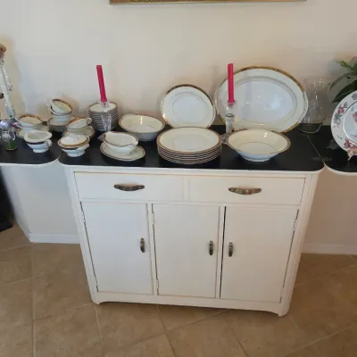 Vintage white-painted wooden buffet cabinet with drop-leaf sides, displaying a gold-rimmed porcelain dinnerware set, decorative plates,...