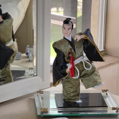 Vintage Japanese samurai doll dressed in a green and gold brocade kimono, displayed on a glass and brass stand in front of a mirror.