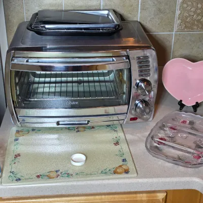 Oster stainless steel toaster oven with baking trays, displayed alongside a pink heart-shaped plate, a glass cutting board, and a divided...