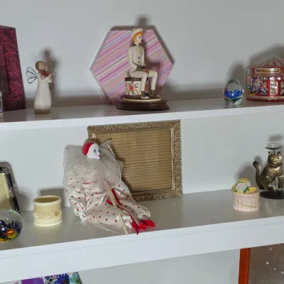 Collection of decorative items on white shelves, including a porcelain clown doll, a Willow Tree angel figurine, picture frames, and...