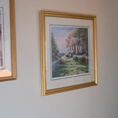 Framed landscape art print depicting a cottage by the sea, matted and housed in a decorative gold-finished frame.