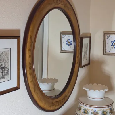 Oval wall mirror with a two-tone wooden frame featuring carved scroll details, displayed alongside framed artwork and a Star of David...