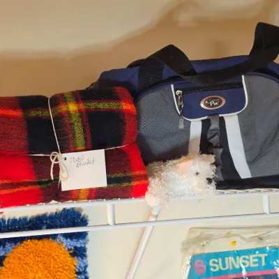 Red and black plaid wool throw blanket tied with string alongside a blue and grey California Pak duffel bag on a wire shelf.
